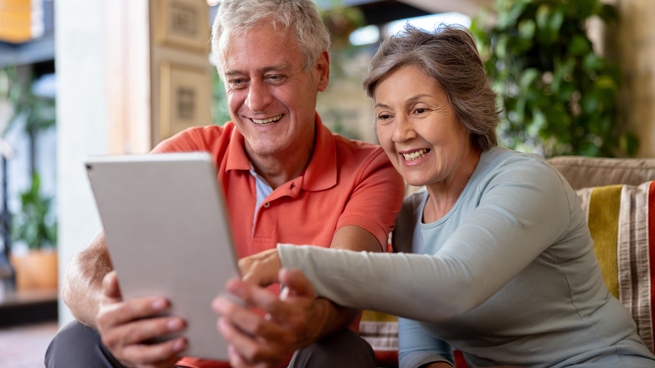 Senior couple smiling while sitting looking and pointing at a tablet.