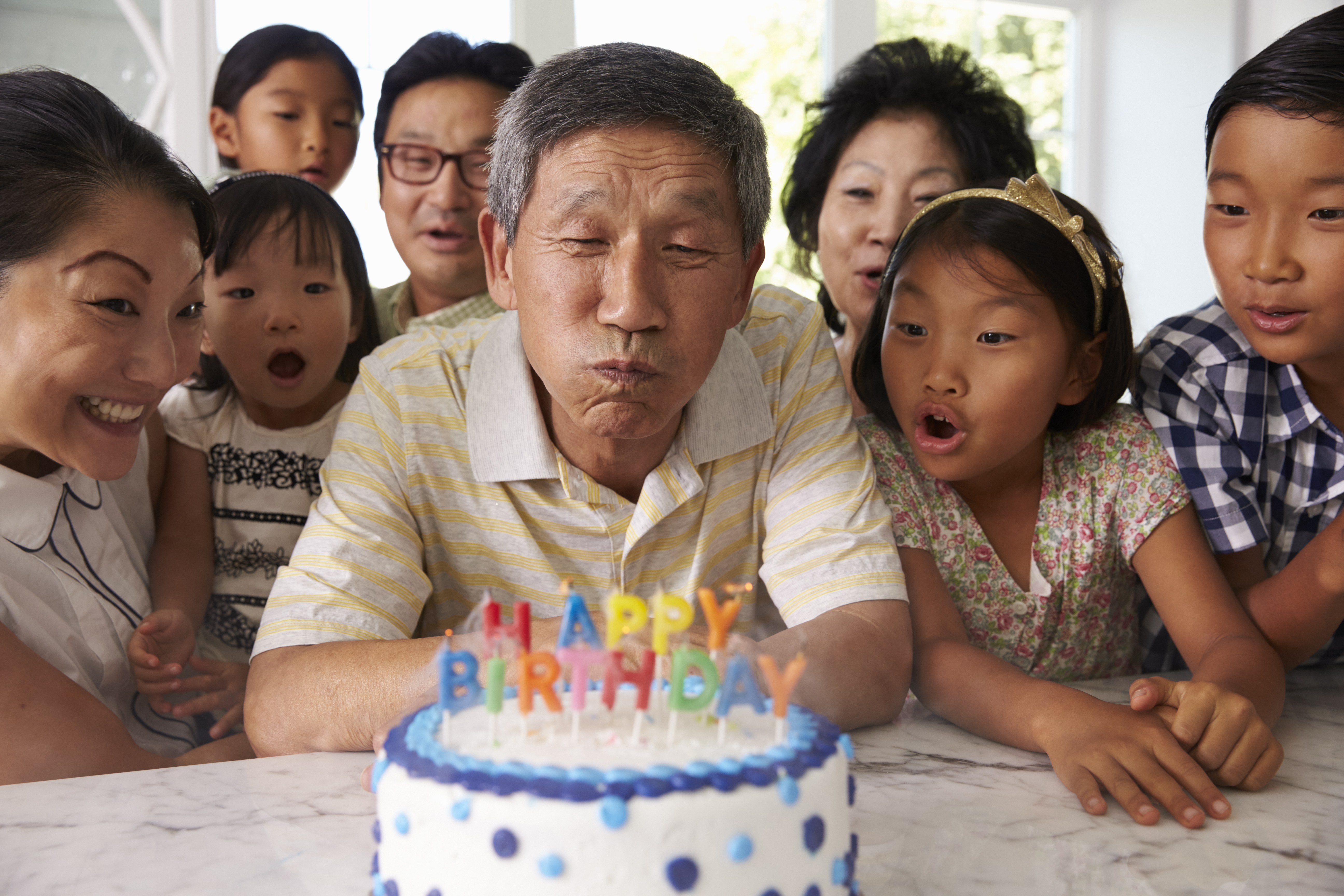 Familia de abuelos, padres y nietos rodeando un pastel y soplando las velas que dicen "Feliz cumpleaños".