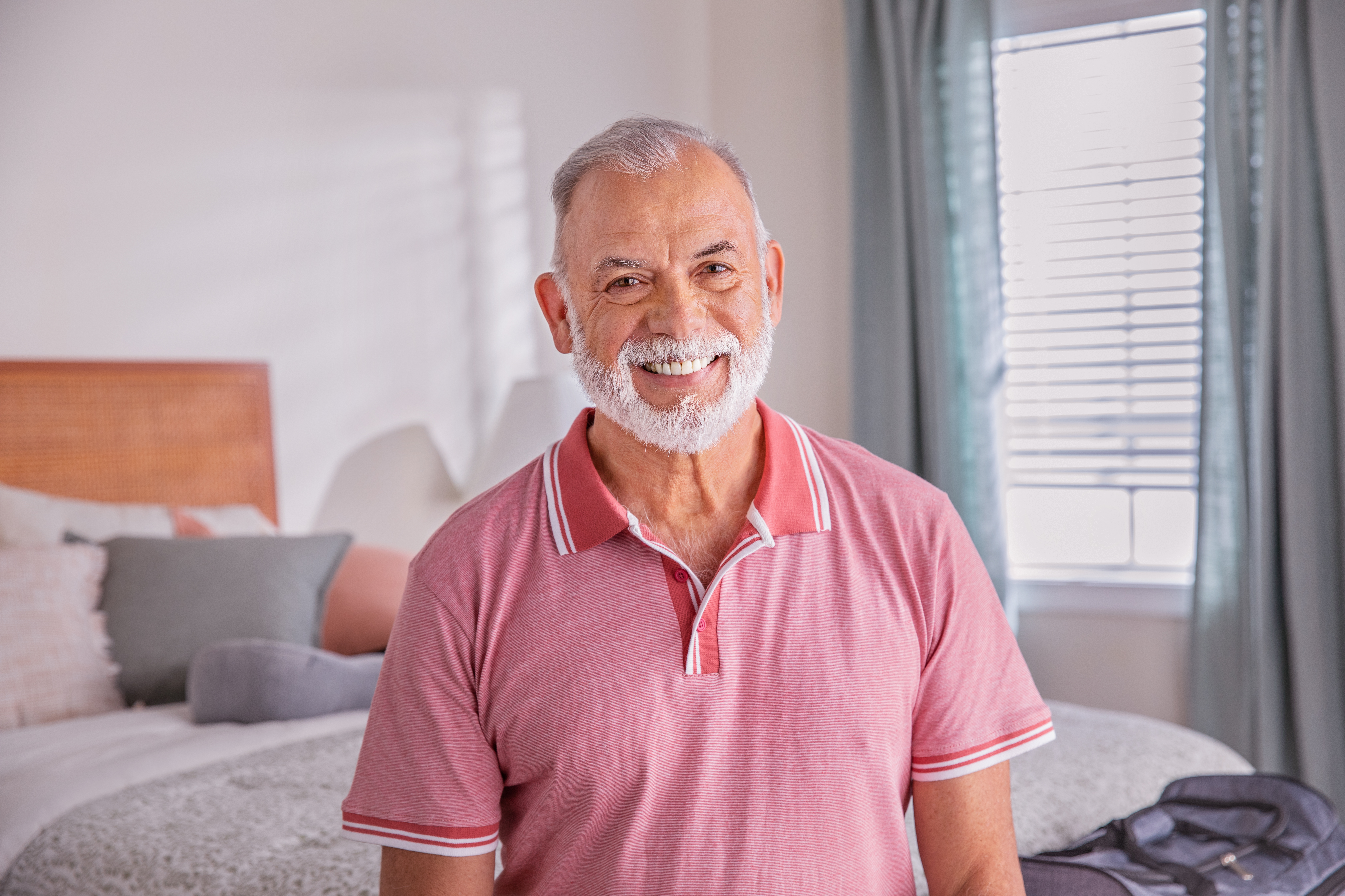 Senior man poses in bedroom sitting on bed next to suitcase