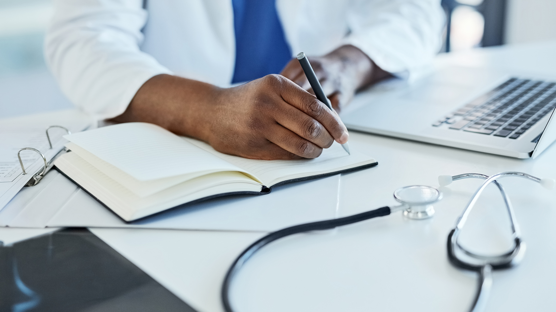 Close-up shot of doctor using a laptop and taking notes.