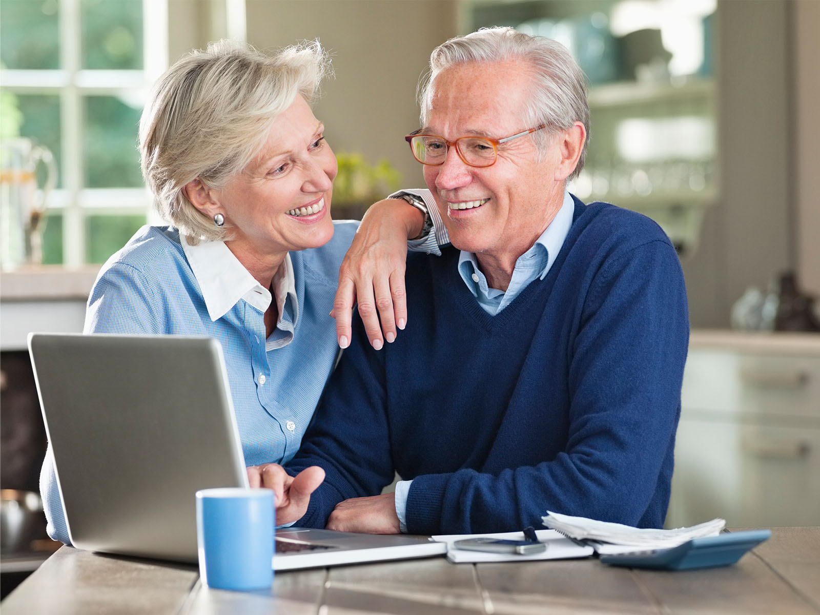 Pareja de personas mayores sonriendo ante una laptop en la mesa de la cocina.