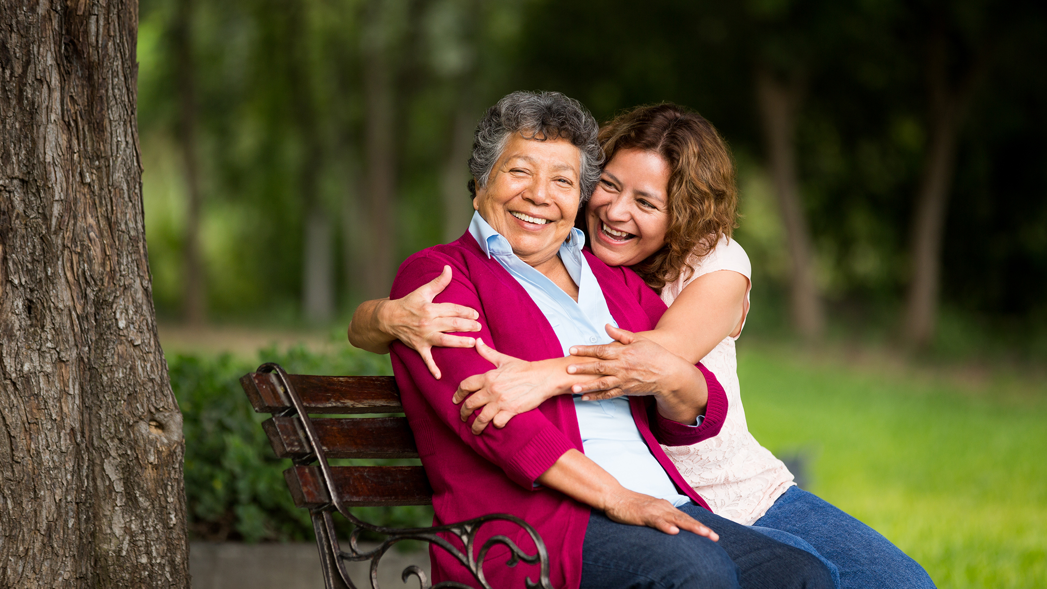 Senior woman being hugged by younger woman on park bench.