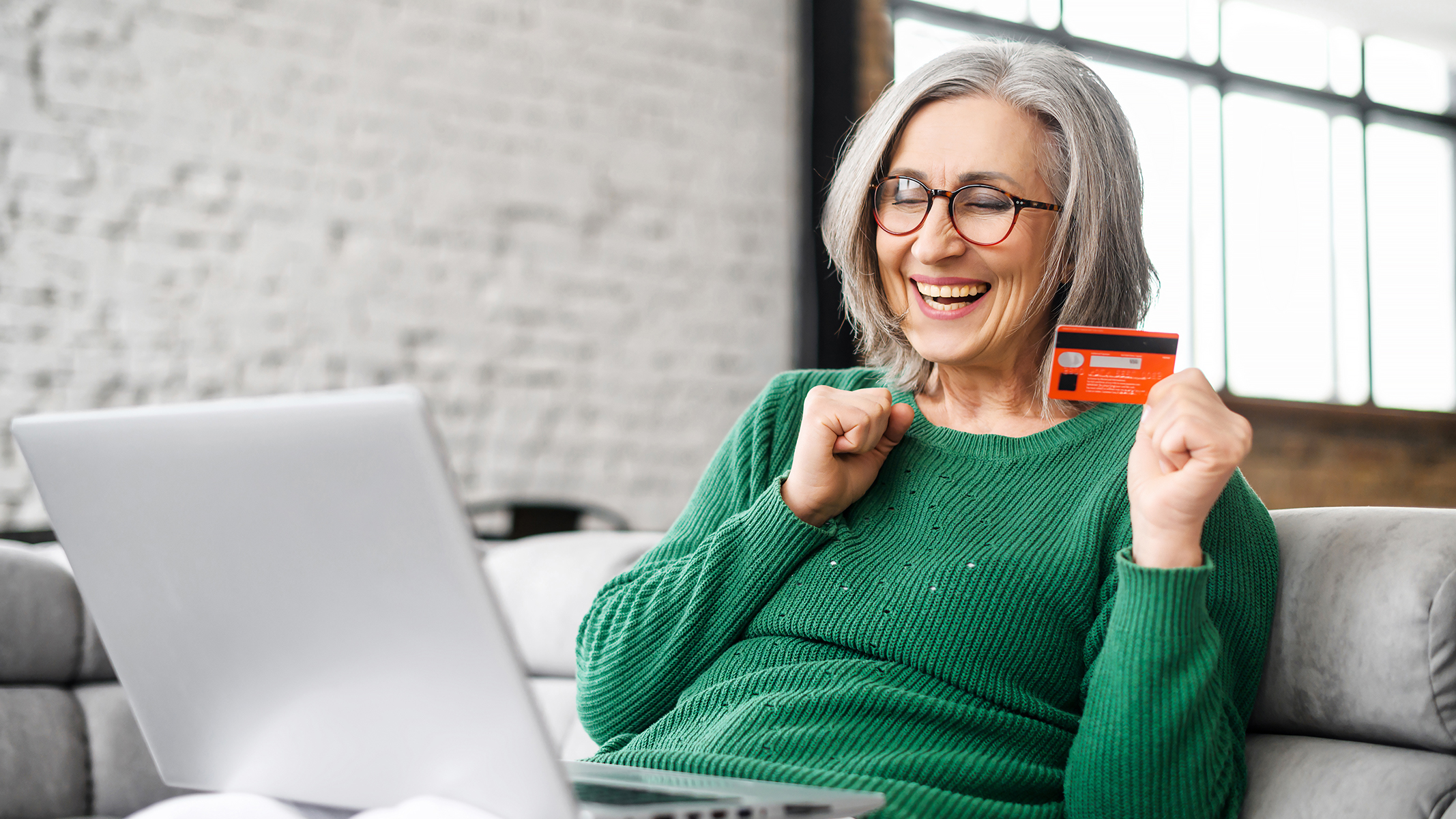 Mujer mayor sonriendo y sentada en el sofá con una tarjeta y una laptop abierta.