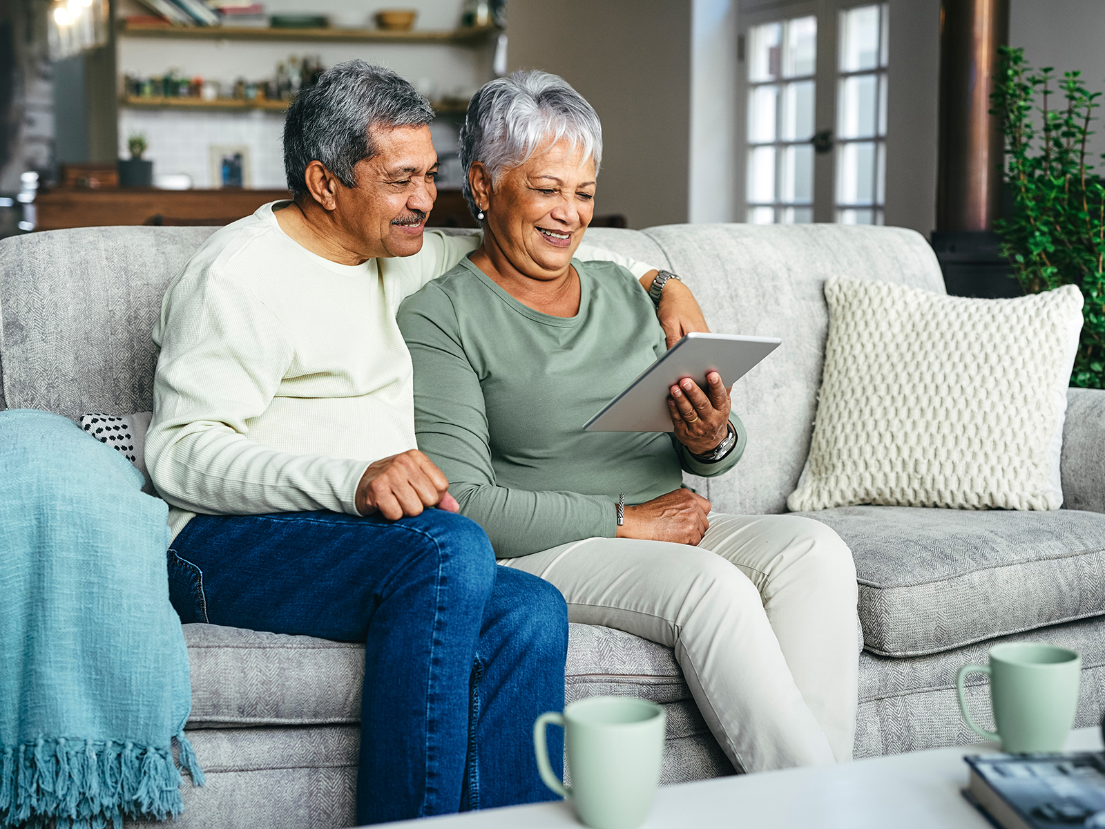 Pareja de personas mayores sonriendo sentados en un sofá mientras miran una tablet.