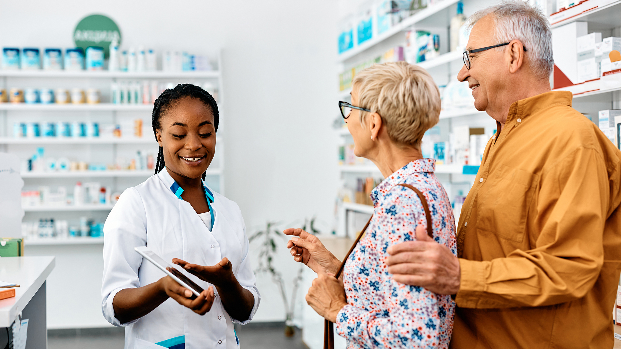 Pareja de personas mayores hablando con una farmacéutica en una farmacia.