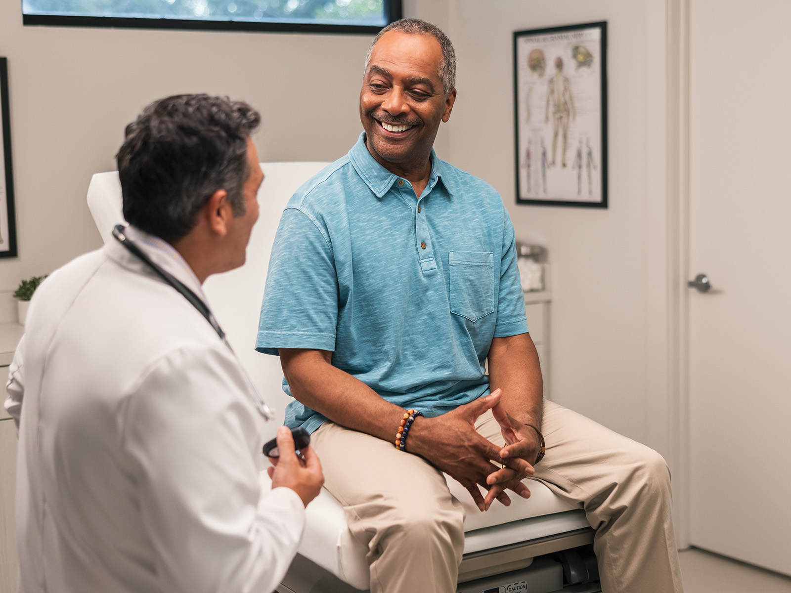  A smiling African-American male patient in a blue shirt and khaki pants is talking with his doctor.