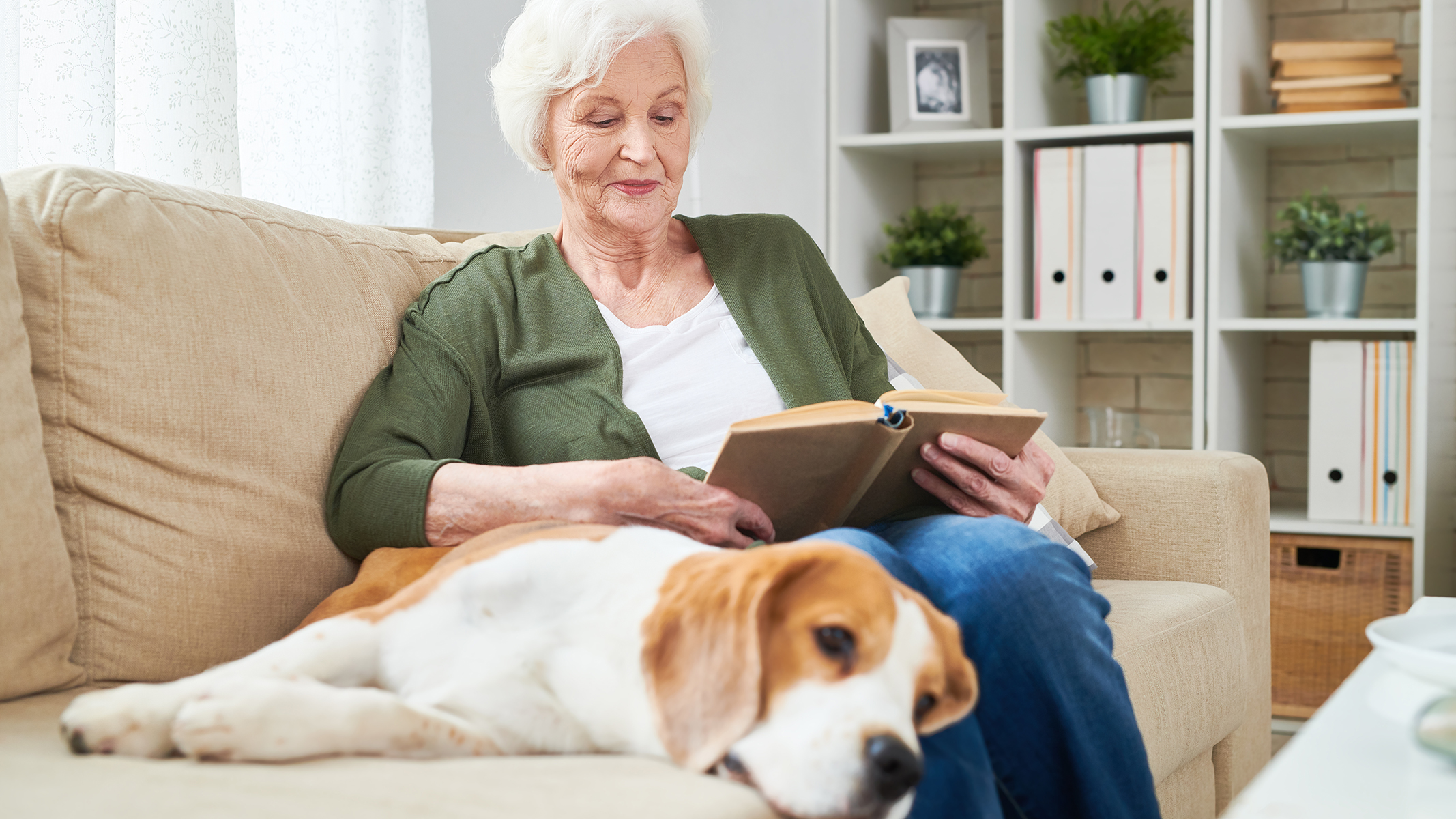 Senior woman reading a book on the couch with a dog.