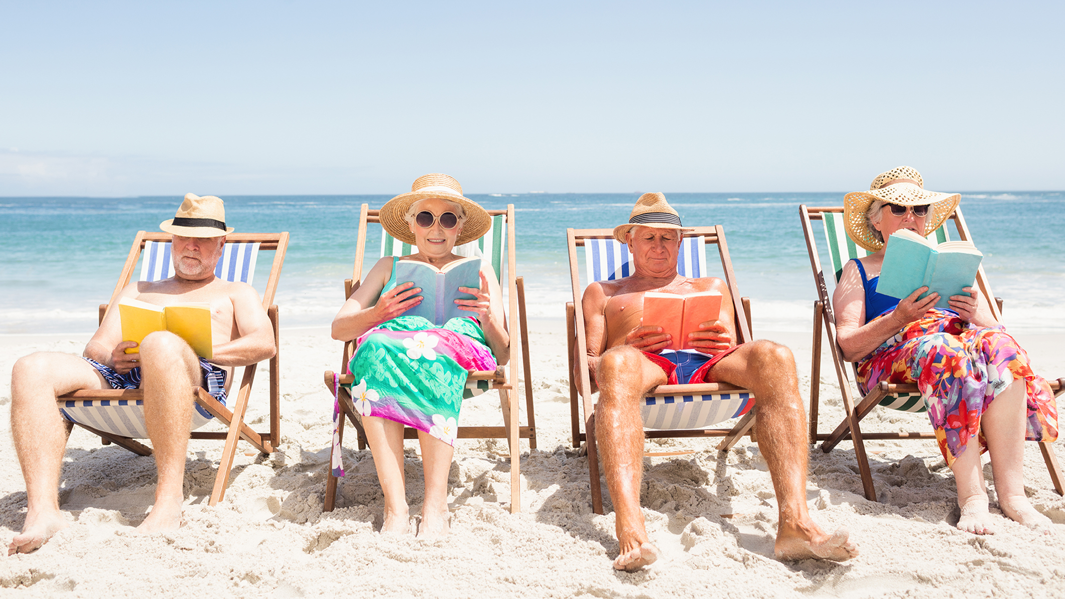 Cuatro personas mayores leyendo libros y descansando en la playa.
