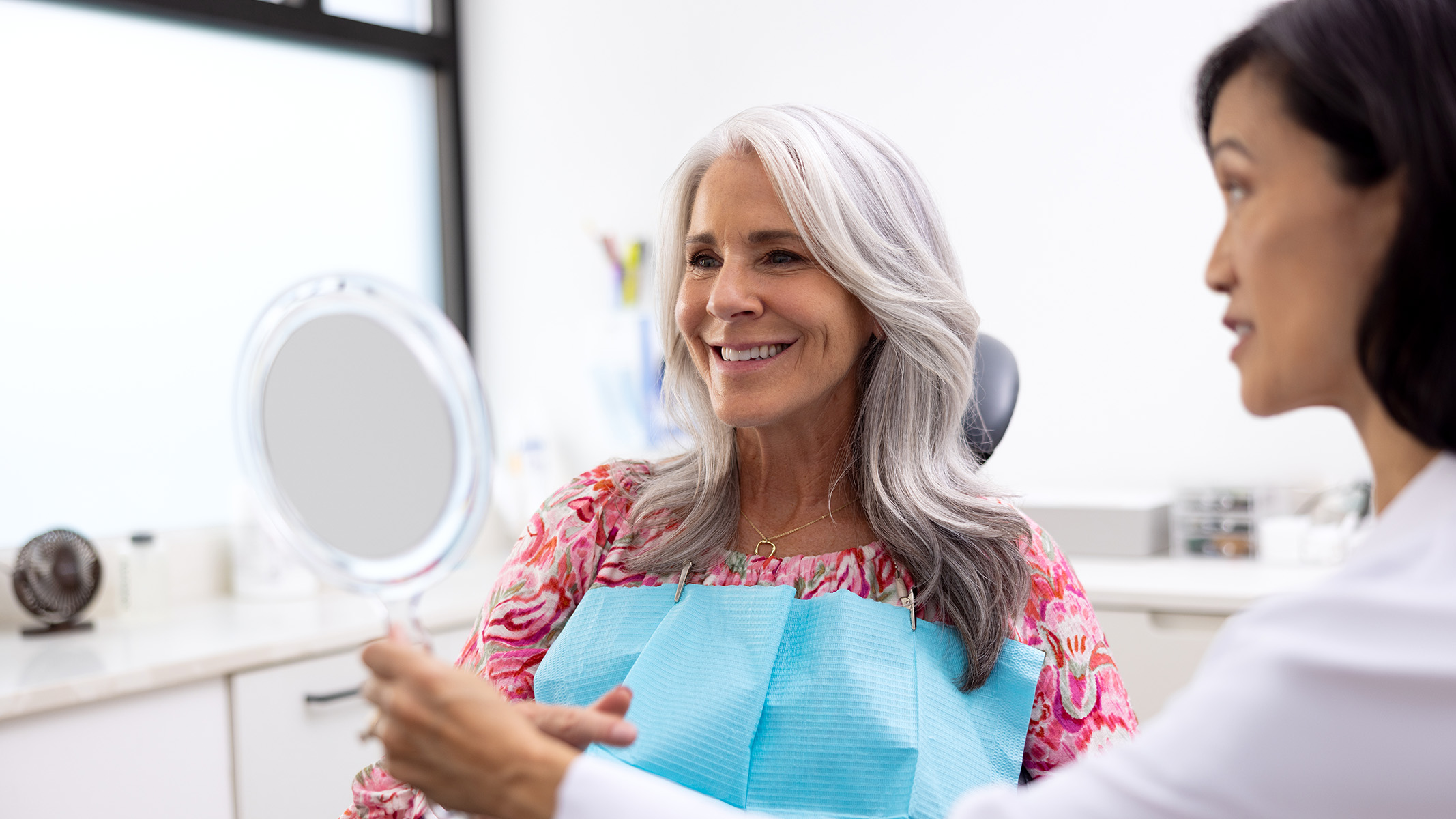 A senior woman smiles into a mirror held by her dentist.